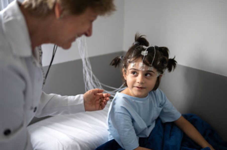 Pediatric EEG setup: a young girl lies in a hospital bed with head electrodes, talking with a clinician nearby.