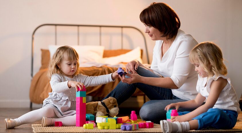 Mother sits on the floor with two young children, playing with colorful building blocks in a cozy bedroom.