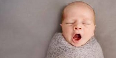 Newborn baby swaddled in a gray blanket, yawning with mouth wide open.