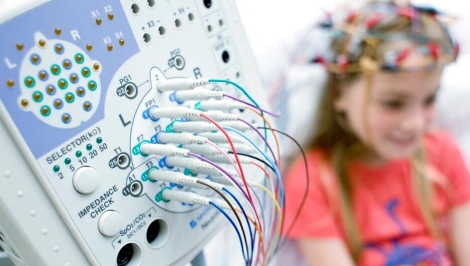 Close-up of a medical device with many colored cables plugged into ports; a smiling child wearing a flower crown sits in the background.