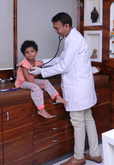Picture3 - Sparsh Hospital - Sparsh Hospital Pediatric doctor in a white coat listening to a young girl's chest with a stethoscope while she sits on a wooden clinic counter.