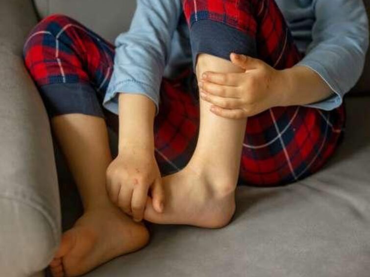 p1 - Sparsh Hospital Child seated on a sofa, holding their ankle with both hands, wearing a blue top and red plaid pajama pants.