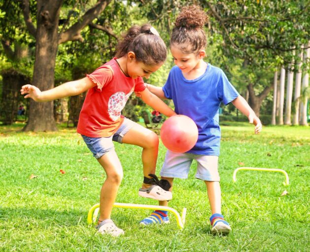p2 - Sparsh Hospital Two young children playing with a pink ball on a grassy park, hopping over small yellow hurdles together