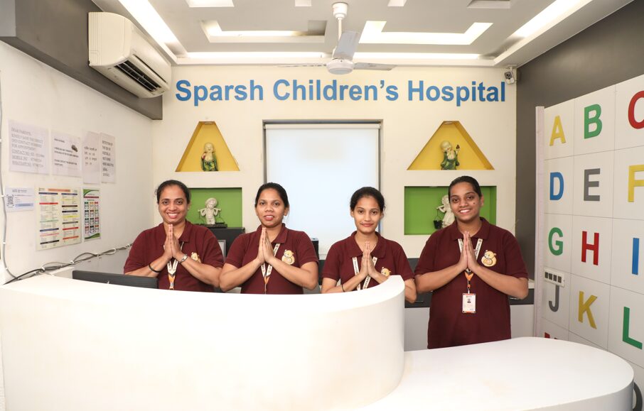 p4 - Sparsh Hospital Reception desk at Sparsh Children's Hospital with four staff members in maroon uniforms greeting visitors with a Namaste gesture.