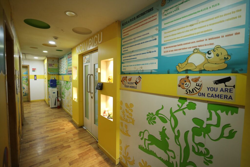Bright hospital hallway with a NICU/PICU entrance, yellow walls, and large informative posters with a cartoon bear on the right wall.