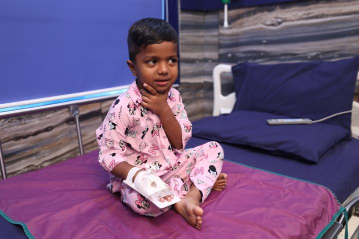 1 - Sparsh Hospital - Sparsh Hospital Young boy in pink pajamas sits on a hospital bed with a bandaged arm and cast, touching his neck.