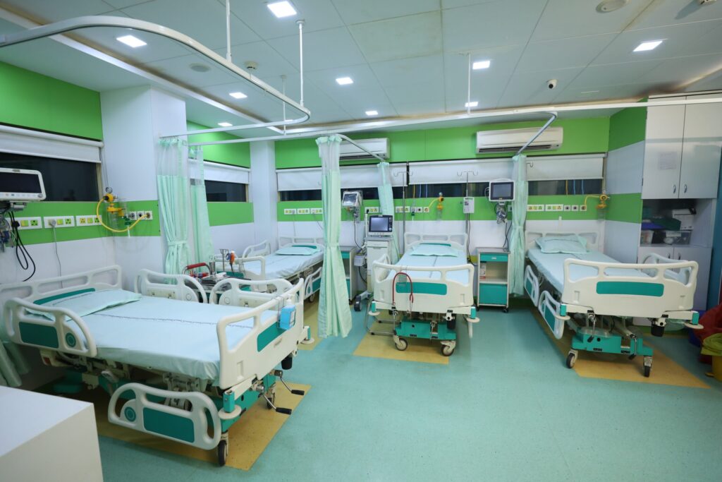 ICU hospital ward with multiple patient beds, green walls, curtains, and monitoring equipment on each bedspace.