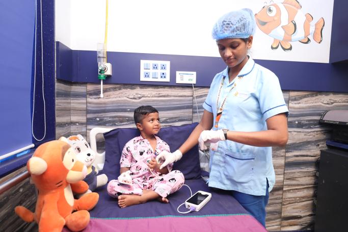 2 - Sparsh Hospital - Sparsh Hospital Pediatric nurse in blue scrubs and a hairnet prepares a procedure for a boy in pink pajamas on a hospital bed; stuffed animals sit nearby as comfort items.