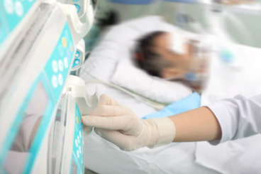 Gloved hand of a clinician adjusting a medical device at a hospital bed with a patient in the background.