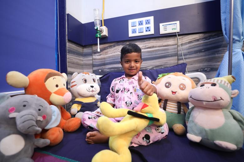 3 - Sparsh Hospital - Sparsh Hospital Smiling child in a hospital bed, pink pajamas, surrounded by stuffed animals, giving a thumbs up in a hospital room.