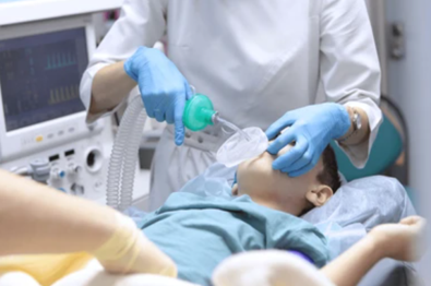 Healthcare professional wearing blue gloves uses a bag-valve mask to ventilate a patient on an operating table, with monitoring equipment in the background.
