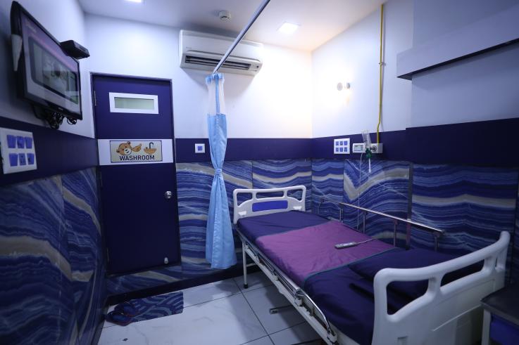 4 - Sparsh Hospital - Sparsh Hospital Hospital patient room with a single bed, purple sheets, blue wave-pattern walls, and a privacy curtain. A wall TV and washroom door are visible.