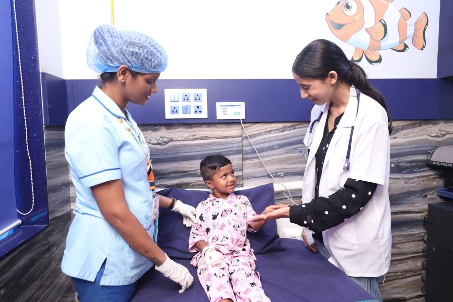 5 - Sparsh Hospital - Sparsh Hospital A young girl in pink pajamas sits on a clinic bed as a doctor and nurse examine her hands in a pediatric room with a fish mural behind them.