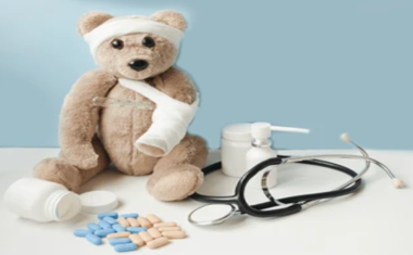 Teddy bear with a white bandage on its arm sits beside a stethoscope, pill bottle, and blue pills on a white table.