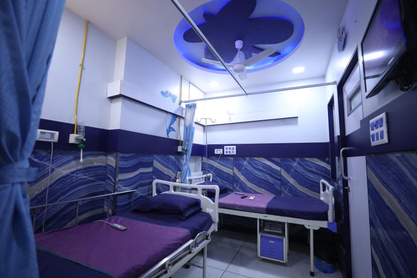 6 - Sparsh Hospital - Sparsh Hospital Hospital ward with two beds, purple linens, blue wavy walls, curtains, and ceiling fan with overhead light