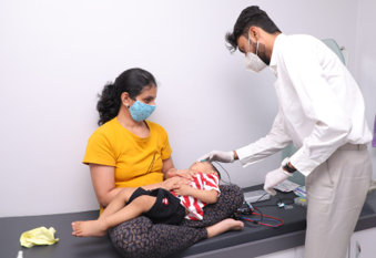 Pediatric exam: a doctor with a mask checks a child on an examination table while a mother sits nearby.