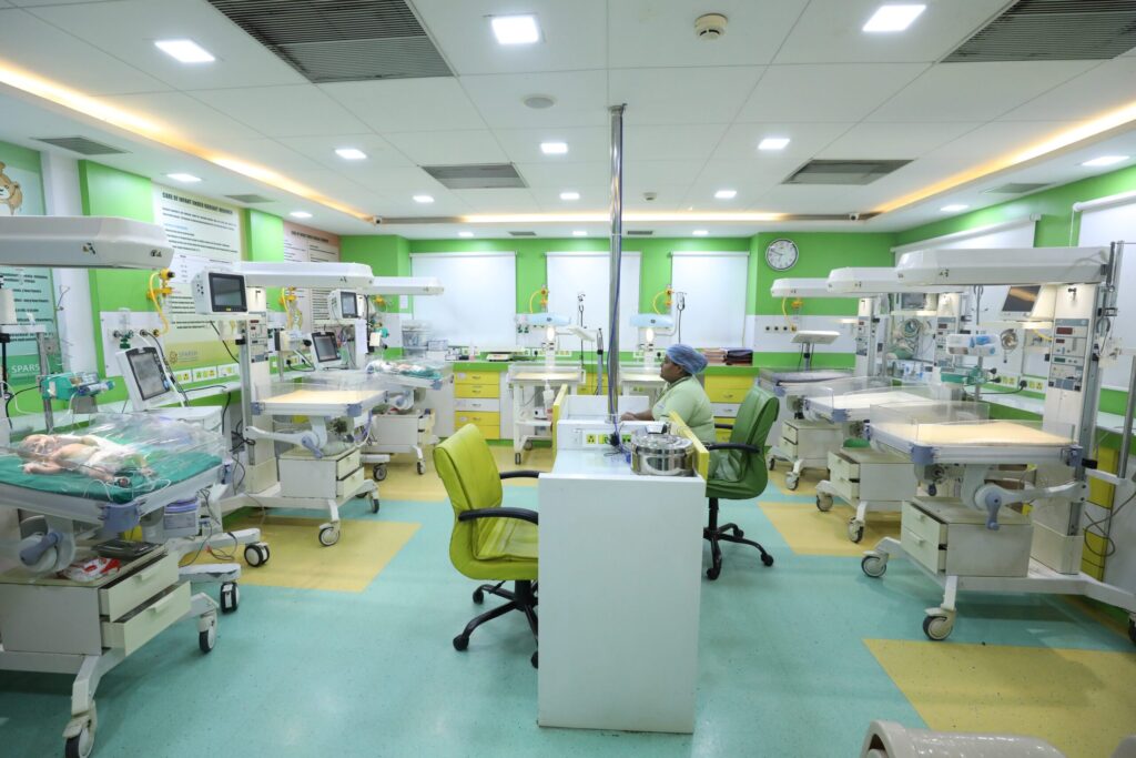 Neonatal intensive care ward with multiple incubators and medical equipment, green walls, and a clinician attending a baby bed.