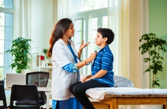 Female doctor with a stethoscope listens to a boy seated on an examination table in a bright clinic room with green walls and plants nearby.