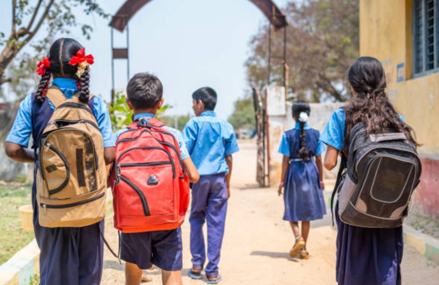Group of students in blue uniforms walking on a dirt path with backpacks toward a school entrance on a sunny day.