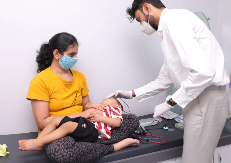 Mother holds her baby on her lap as a clinician in a white coat and mask examines the child at a clinic counter in a pediatric checkup setting.