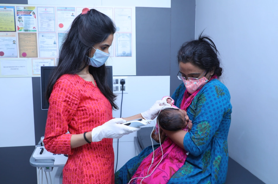 Nurse wearing a mask and gloves uses a medical device on a newborn held by a mother in a clinic.