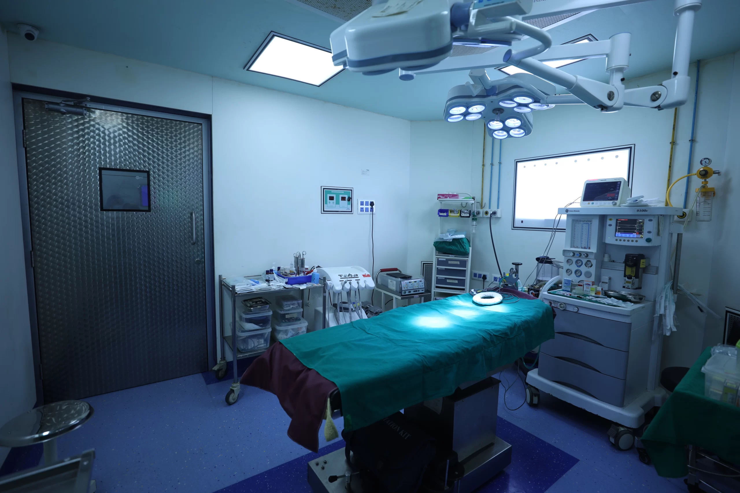 Operating room with a patient bed draped in teal, overhead surgical lights, and medical monitors nearby.