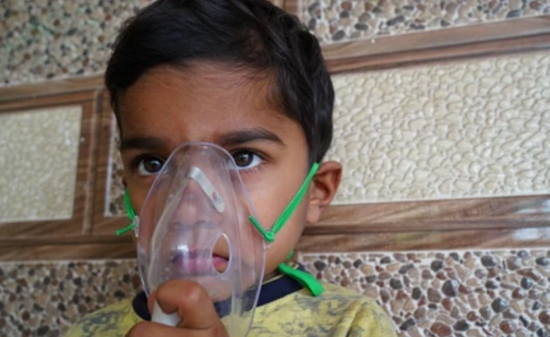 Breating Problem - Sparsh Hospital - Sparsh Hospital Young boy wearing a clear medical inhalation mask with green straps, sitting on tiled stairs.