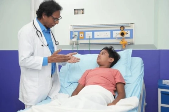 Pediatric Inpatient Care for Adolscent - Sparsh Hospital - Sparsh Hospital Doctor in a white coat with a stethoscope speaks softly to a boy lying in a hospital bed beside blue curtains.
