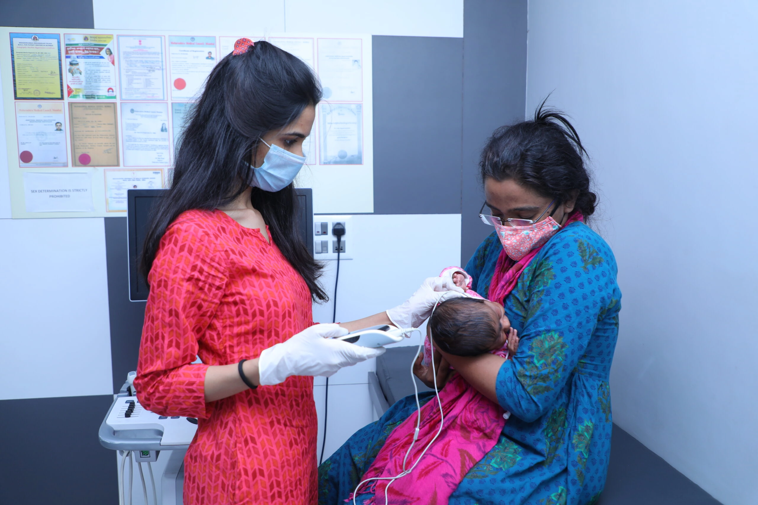 Pediatric Inpatient Care for new born - Sparsh Hospital - Sparsh Hospital Nurse in a red patterned dress, wearing a mask and gloves, using a medical device on a newborn held by a mother in a blue sari, inside a clinic.