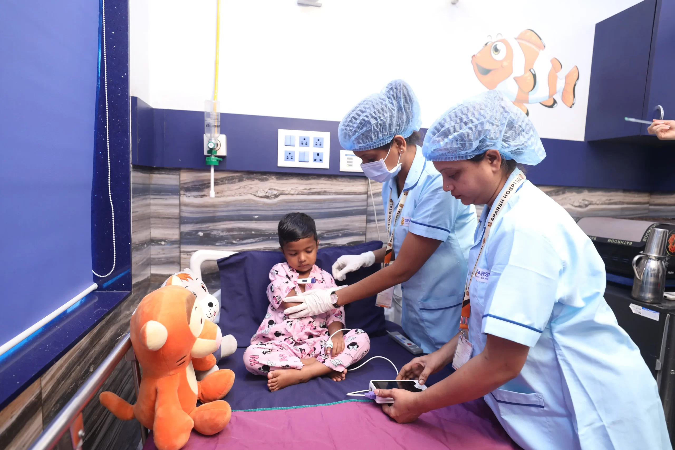 Services Included in Inpatient Pediatric - Sparsh Hospital - Sparsh Hospital Two nurses in blue scrubs with surgical caps and masks assist a young patient in a hospital bed, with plush tiger toys nearby.