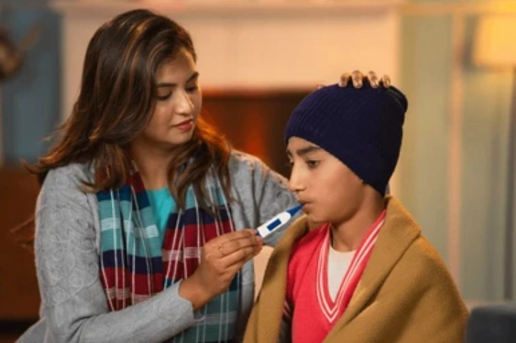 Severe Infection - Sparsh Hospital - Sparsh Hospital Woman checks a boy's temperature with a thermometer while comforting him with a hand on his shoulder and a warm scarf nearby.