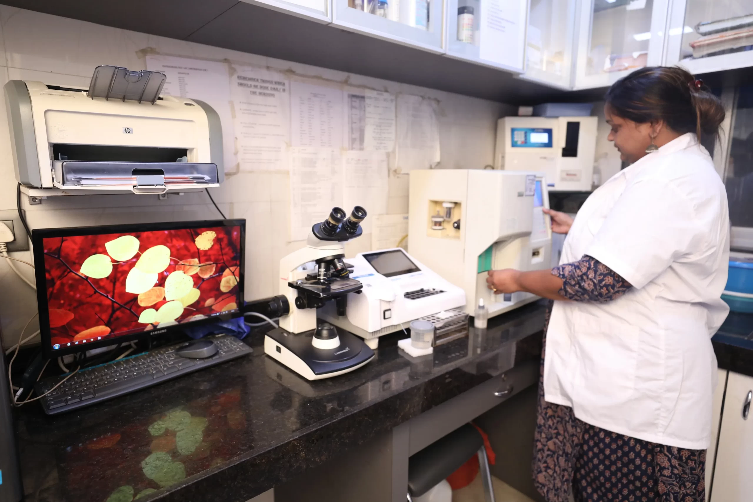 Why Pediatric Laboratory services are important - Sparsh Hospital - Sparsh Hospital Female lab technician in a white lab coat stands at a cluttered benchtop, using a microscope with a computer monitor showing a colorful leaf image nearby.