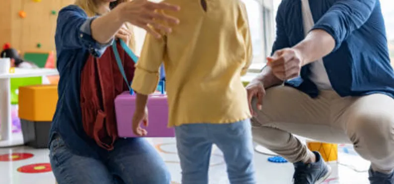 Two adults kneel beside a toddler in a bright playroom; one reaches toward the child, the other holds a purple toy box.