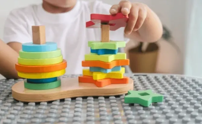 Child stacks colorful wooden rings onto a base, building a tower with a toy on a table.