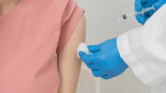 Vaccines for Toddlers and Older Children - Sparsh Hospital - Sparsh Hospital Person in a pink shirt receives a vaccination in the upper arm from a clinician wearing blue gloves visible with a syringe nearby.