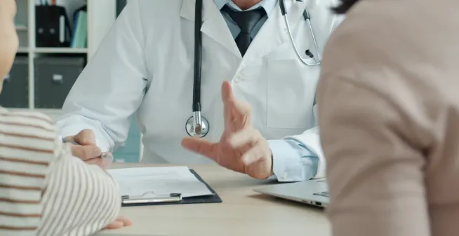Doctor in a white coat with a stethoscope consults a patient across a desk, with a clipboard and laptop nearby.