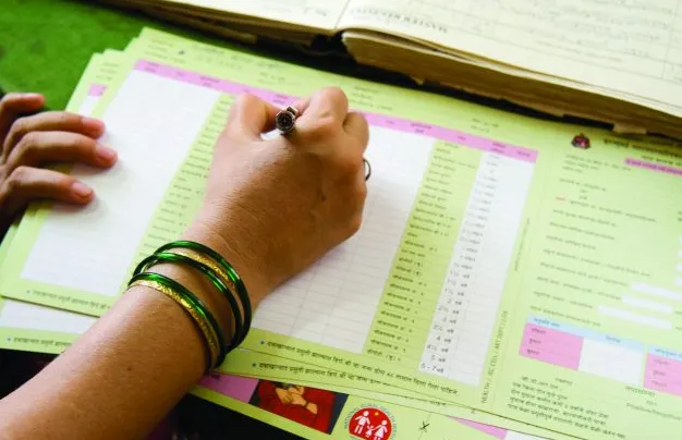 What to Bring for a Vaccination Visit - Sparsh Hospital - Sparsh Hospital Person writes with a pen on a bright green form or chart, wearing green bracelets on their wrist.
