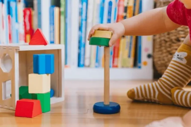 Child stacks a block on a wooden tower while colorful blocks sit nearby on a light wooden floor.