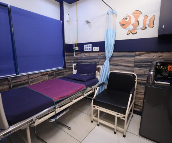 Hospital room with an adjustable bed, blue privacy curtain, and a small seating chair beside a kitchenette with a mini fridge and microwave; wall mural shows a clownfish.