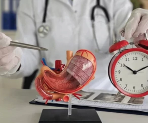 Clinician in white coat and gloves holds a surgical instrument beside a realistic heart model and a red alarm clock on a desk. (teaching/medical demo)