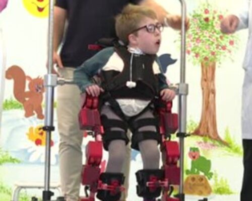 Child in leg braces (gait orthosis) standing in a supportive walker, held by a caregiver, with a colorful mural backdrop.