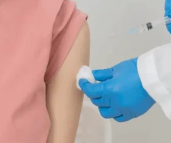 Person in a pink shirt receives a vaccination in the upper arm from a clinician wearing blue gloves visible with a syringe nearby.