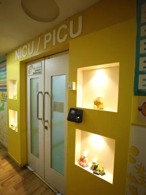 NICU/PICU entrance in a yellow hall: white double doors with glass, 'NICU/PICU' sign above, and small lit display shelves.